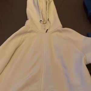 Vit hoodie med dragkedja från Peak Performance - Säljer en stilren vit hoodie från Peak Performance med dragkedja framtill och huva med snörning. Tröjan har broderad logga på bröstet och är perfekt för en avslappnad look.