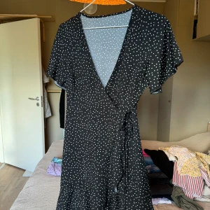 Svart omlottklänning med vita prickar - Svart omlottklänning med vita prickar och knyt i midjan. Klänningen har korta ärmar och v-ringad halsringning.