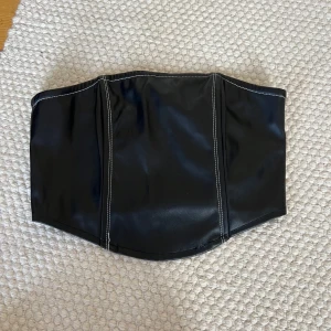 Svart topp i skinnimitation - Säljer en svart tubtopp i skinnimitation med markerade vita sömmar. Toppen har dragkedja bak. Helt oanvänd med lappar kvar 