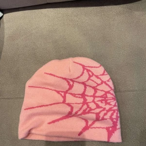 Rosa mössa med spindelnätsmönster - Cool rosa mössa med ett iögonfallande spindelnätsmönster i mörkare rosa. Tillverkad i 100% akryl, mjuk och stretchig passform. Perfekt accessoar för att sticka ut i mängden.