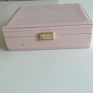 Ljusrosa smyckeskrin med gulddetalj😍 - Säljer ett elegant ljusrosa smyckeskrin med guldfärgad låsdetalj. Skrinet har flera fack och ringrader för smidig förvaring av smycken som ringar, halsband och armband. Insidan är klädd i mjukt tyg och har elastiska fickor och krokar för extra organisering.