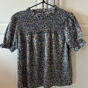 Blommig blus med puffärm - Supersöt blommig blus i blått, beige och lila med puffärmar och rynkad överdel. Blusen har liten volang vid halsen och knapp i nacken. Perfekt för vår och sommar!
