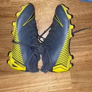 Nike Mercurial fotbollsskor i grått och gult - Säljer ett par Nike Mercurial fotbollsskor i grått med gula detaljer och svart Nike-logga. Skorna har snörning och dobbar för spel på gräs. Snygg sportig design med hög komfort och bra grepp.