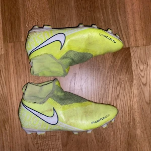 Gula Nike Phantom Vision fotbollsskor - Säljer ett par gula Nike Phantom Vision fotbollsskor med vit och svart swoosh. Skorna har en strumpliknande passform utan traditionell snörning och är designade för spel på gräs. Perfekta för dig som vill sticka ut på planen!