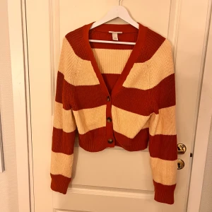 Randig kofta från Monki - Säljer en stickad kofta från Monki i storlek XS. Koftan har breda ränder i rostbrunt och beige, knappar framtill och är croppad i modellen. Perfekt för lager-på-lager och snygg till både jeans och kjol.