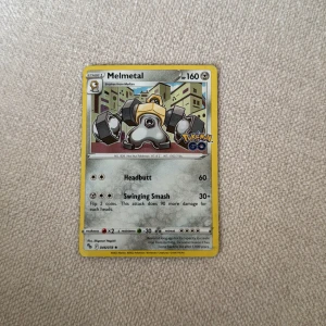 Pokémonkort Melmetal - Säljer ett Pokémonkort med Melmetal från Pokémon GO-serien. Kortet har gul ram och visar Melmetal i en stadsmiljö. HP 160 och attacker Headbutt samt Swinging Smash. Baksidan har klassisk blå design med Pokéboll.