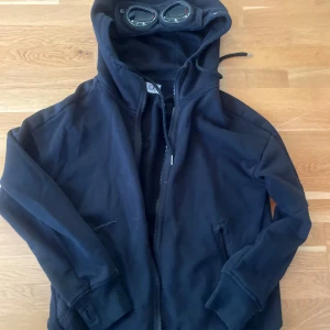 Svart hoodiejacka med glasögon i huvan från C.P. Company - Säljer en svart hoodiejacka från C.P. Company med dragkedja och unika glasögon integrerade i huvan. Jackan har två fickor framtill och ribbade muddar vid ärmslut och nederkant. Perfekt för dig som vill sticka ut!