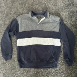 Randig pikétröja i blått och grått från Murrey  - Säljer en klassisk pikétröja från Murrey med breda ränder i mörkblått och två nyanser av grått. Tröjan har lång ärm, krage och knäppning vid halsen. Perfekt för en avslappnad stil.