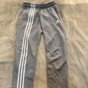 Grå mjukisbyxor från Adidas med vita ränder - Säljer ett par grå mjukisbyxor från Adidas med klassiska vita ränder längs ena benet och Adidas-logga på låret. Byxorna har dragsko i midjan och är perfekta för en avslappnad stil.