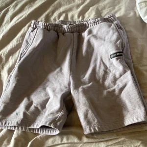 Ljuslila shorts från Nicki Studios - Säljer ett par ljuslila shorts från Nicki Studios med resår i midjan och sidofickor. Mjukt material och enkel design med en svart logotyp-patch på vänster ben. Perfekta för chill dagar eller när du vill vara bekväm. OBS! finns färgfläckar