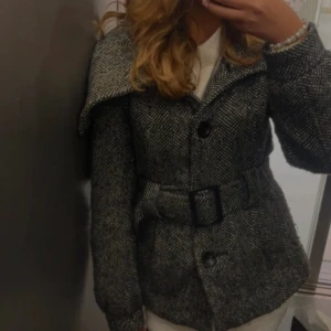 Grå kappa med bälte - Säljer en snygg grå kappa med bälte i midjan och stora knappar framtill. Jackan har lång ärm och bred krage, perfekt för kyligare dagar. Klassisk och tidlös modell som passar till många olika stilar.