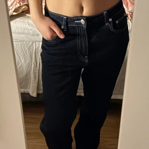 Mörkblå jeansbyxor från NA-KD - Säljer ett par mörkblå jeansbyxor från NA-KD med kontrasterande vita sömmar. Byxorna har klassisk femficksdesign, raka ben och hög midja. Perfekta för dig som gillar en stilren och enkel look.