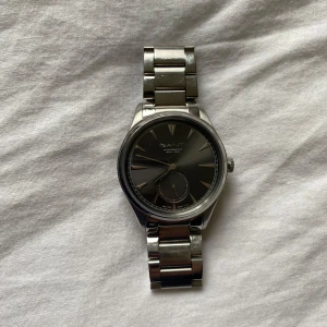 Herrklocka GANT - Stilren klocka från GANT med svart urtavla och silverfärgat armband i metall. Klockan har minimalistisk design med tydliga index och en extra liten sekundvisare. Perfekt accessoar för dig som gillar klassisk stil.