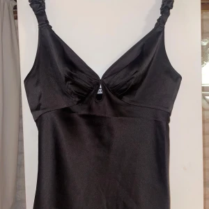 Svart satinlinne med rynkade axelband från Adoore - Snygg svart linnetopp i satin från Adoore med rynkade axelband och liten öppning framtill. Perfekt för dig som gillar stilrena och eleganta toppar med en twist. Säljes i storlek 42. 