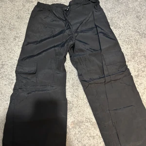 Svarta cargobyxor från Missy Empire - Säljer ett par svarta cargobyxor från Missy Empire med dragsko i midjan och stora fickor på sidorna. Byxorna har en loose passform och är perfekta för en avslappnad stil. Materialet känns lätt och syntetiskt.