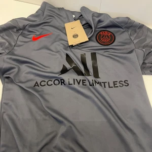 Grå PSG fotbollströja från Nike - Säljer en grå Paris Saint-Germain fotbollströja från Nike med röd logga och svart PSG-märke på bröstet. Tröjan har svart tryck med 'Accor Live Limitless' på framsidan och är tillverkad i ett lätt, andningsbart material.Helt nytt skick skriv om du vill få reda på nån annan info eller mer bilder. Om du är intresserad av dess byxor skriv de