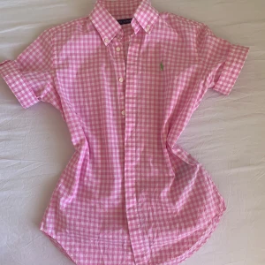Rosa rutig skjorta från Ralph Lauren - Säljer en rosa och vit rutig skjorta från Ralph Lauren med korta ärmar och klassisk krage. Skjortan har knappar framtill och en grön broderad logga på bröstet. Perfekt för en fräsch och preppy stil!