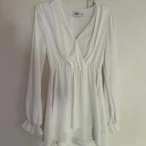 Vit klänning  - Supersöt vit klänning med v-ringning och långa ärmar. Klänningen har resår i midjan och volangdetalj nertill, vilket ger en flowy känsla.