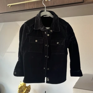 Svart manchesterjacka från KappAhl - Säljer en svart manchesterjacka från KappAhl med klassisk krage och knäppning framtill. Jackan har två bröstfickor med lock och knappar samt knappar vid ärmsluten. Perfekt för dig som gillar en tidlös stil.
