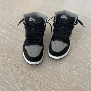 Nike Air Jordan 1 High svart och grå - Säljer ett par Nike Air Jordan 1 High i svart och grå med vit sula. Skorna har snörning, rund tå och är tillverkade i skinn med detaljer i syntet. Klassisk Jordan-logga på sidan och Nike Air-märke på plösen.