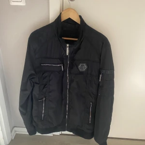 Svart jacka från Philipp Plein äkta köpt för 3 år sen - Säljer en svart jacka från Philipp Plein med silverfärgade dragkedjor och logotypdetalj på bröstet. Jackan har flera fickor med dragkedja och en markerad logotyp på ärmen. Snygg och stilren design med hög krage.