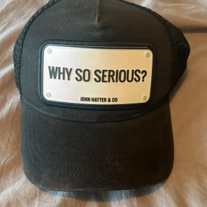 Svart keps från John Hatter & Co - Svart keps med mesh-bakdel och justerbar passform. Framtill sitter en vit skylt med texten 'WHY SO SERIOUS?'. Märket John Hatter & Co är tydligt markerat. Perfekt för en cool och avslappnad stil.