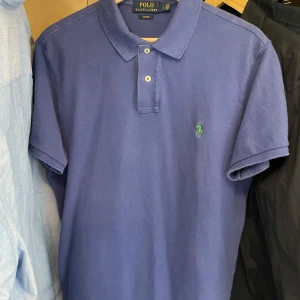 Blå pikétröja från Polo Ralph Lauren - Klassisk blå pikétröja från Polo Ralph Lauren med korta ärmar och krage. Tröjan har två knappar framtill. Perfekt för en stilren och avslappnad look.