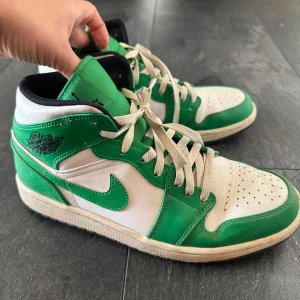 Nike Air Jordan 1 Mid vit och grön - Säljer ett par Nike Air Jordan 1 Mid i vit och grön färg. Skorna har klassisk snörning, rund tå och platt sula. Ikonisk Jordan-logga på sidan och tungan. Perfekta för dig som gillar streetwear och sneakers med retrostil.