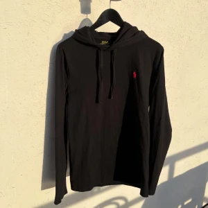 Svart hoodie från Ralph Lauren - Svart tunn hoodie från Polo Ralph Lauren med klassisk huva och dragsko. Tröjan har lång ärm och en liten röd broderad logga på bröstet. Perfekt för en avslappnad och stilren look.