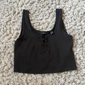 Svart croppad linnetopp från Vero Moda - Svart ribbad croppad linnetopp från Vero Moda med snygg snörning framtill. Toppen är ärmlös och har en djup rundad urringning. Perfekt att matcha med jeans eller kjol för en trendig look.