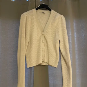 Beige ribbad kofta med knappar - Säljer en beige ribbad kofta med v-ringning och stora knappar framtill. Tröjan har lång ärm och är lite croppad i modellen. Perfekt att slänga över en topp för en enkel men snygg look.