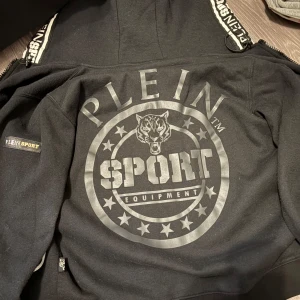 Plein sport hoodie/jacka - Svart hoodie med stor logga och vargmotiv på ryggen från Plein Sport. Huva med dragsko och dragkedja, samt tryckta detaljer på ärmarna. Perfekt för en sportig och street-inspirerad stil.