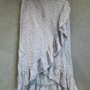 Ljuslila blommig omlottkjol med volang - Säljer en ljuslila långkjol med småblommigt mönster och omlott-design. Kjolen har volangdetaljer längs kanten och nertill, vilket ger en romantisk och flowy känsla. Perfekt för dig som gillar en feminin stil.