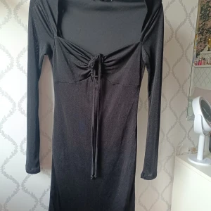 Svart långärmad klänning  - Säljer en svart långärmad klänning från Bik Bok med snygg knytning framtill vid bysten. Klänningen har en figurnära passform och är tillverkad i ett mjukt, stretchigt material. Perfekt för dig som gillar stilrena och enkla plagg. Helt Nya och aldrig använd!!! Nypris 399,mitt pris 279