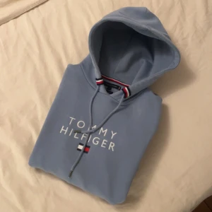 Ljusblå hoodie från Tommy Hilfiger - Säljer en ljusblå hoodie från Tommy Hilfiger med broderad logga och klassiska färgdetaljer vid huvan. Tröjan har dragsko i huvan och är perfekt för en avslappnad stil. passar S ! 