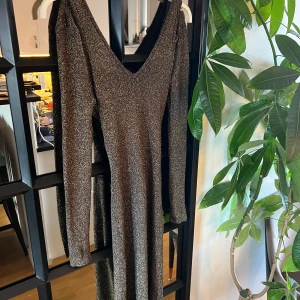 Glittrig långklänning med cut out-detalj - Säljer en svart långklänning med glittrigt material och v-ringning. Klänningen har långa ärmar och en snygg cut out-detalj framtill. Perfekt för dig som vill sticka ut med en elegant och trendig look.