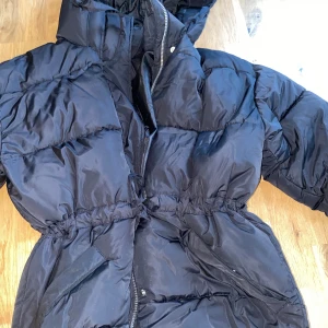 Svart pufferjacka med huva - Säljer en svart pufferjacka med huva och dragkedja. Jackan har lång ärm, markerad midja och är vadderad för extra värme. Perfekt för kyliga dagar och har en sportig look.