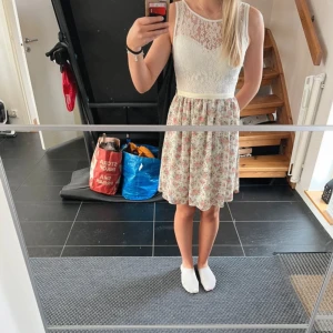 Vit och blommig ärmlös klänning med spets - Superfin ärmlös klänning med vit spets upptill och blommigt mönster nertill. Klänningen har rund halsringning, markerad midja med band och kjolen är lätt utställd. Perfekt för vår och sommar.