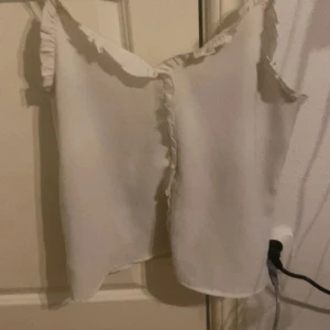 Vit volangprydd linnetopp - Säljer en söt vit linnetopp med volangdetaljer längs axelbanden och framtill. Toppen är ärmlös och har en luftig passform, perfekt för varma dagar.