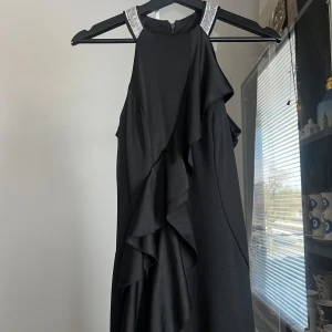 Svart långklänning med volang och glittriga axelband - Säljer en svart långklänning med höghalsad design och dekorativa volanger framtill. Klänningen har glittriga axelband som ger en extra festlig touch. Perfekt för speciella tillfällen när du vill sticka ut. (Använt en gång )