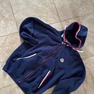 Marinblå hoodie från Moncler i bra skick lite sliten men fortfarande i ganska bra skick diskutera pris i dm! - Säljer en marinblå hoodie från Moncler med dragkedja och huva. Tröjan har vita och röda detaljer längs dragkedjan och fickorna samt Moncler-logga på bröstet. Perfekt för en sportig och stilren look.