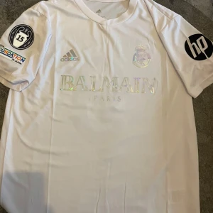 Vit Real Madrid x Balmain fotbollströja Mbappé 9 - Snygg vit fotbollströja från Adidas i samarbete med Balmain och Real Madrid. Tryck med Mbappé och nummer 9 på ryggen, samt flera loggor på ärmarna. Klassisk passform och korta ärmar. Perfekt för fotbollsfans!