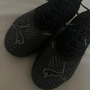 Svarta fotbollsskor från Puma - Säljer ett par svarta fotbollsskor från Puma med vit logga och tunna linjer på ovandelen. Skorna har snörning och dobbar i mintgrönt för bra grepp på planen. Perfekta för dig som vill ha coola och snygga fotbollsskor. Passar både tjejer och killar storlek 40,5 men sitter bra på 40 och 39,5. Har använt den 1 gång och nu spelar jag inte fotboll längre, pris kan diskuteras💙🤍