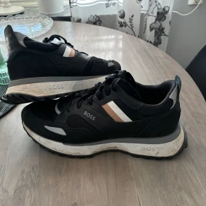 Svarta sneakers från BOSS - Säljer ett par svarta sneakers från BOSS med vita och beige detaljer på sidan. Skorna har snörning, rund tå och platt sula. Materialmix av skinn och syntet med meshpartier för extra komfort. Perfekta för en stilren och sportig look.