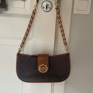 Brun axelväska från Michael Kors - Säljer en snygg brun axelväska från Michael Kors med kedjerem och logga i guld framtill. Väskan har ett klassiskt MK-mönster, detaljer i brunt skinn och innerfack med dragkedja. Perfekt för dig som vill ha en stilren accessoar.