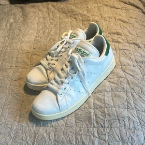Adidas Stan Smith vita sneakers med gröna detaljer - Klassiska vita Adidas Stan Smith sneakers med gröna detaljer på hälen och tungan. Skorna har snörning, platt sula och rund tå. Perfekta för en stilren och sportig look. Adidas-logga syns tydligt bak och på plösen.