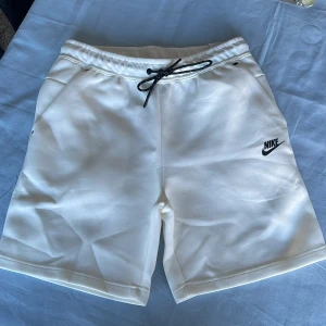 Vita shorts från Nike med snörning - Säljer ett par vita Nike tech shorts med svarta snören. Shortsen har fickor på sidorna och är tillverkade i ett mjukt material som passar perfekt för träning eller varma dagar. Ganska liten i storleken så skulle rekomendera om du har M. Aldrig använda