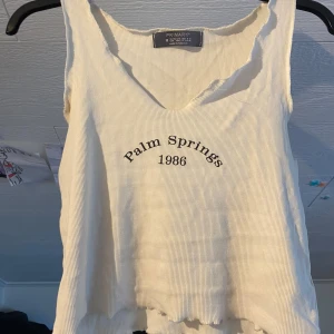 Vit linnetopp från Primark - Säljer en vit linnetopp från Primark med texten 'Palm Springs 1986' framtill. Toppen har v-ringning och är ärmlös, perfekt för varma dagar. Enkel och stilren design som passar till mycket.