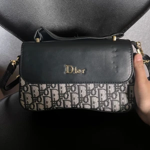 Svart och beige axelväska - Snygg axelväska (inte äkta!), svart skinn med beige och svart monogrammönster. Väskan har guldfärgade detaljer, lock med fin logga och justerbar axelrem. Perfekt för att lyfta din outfit med en fin touch.