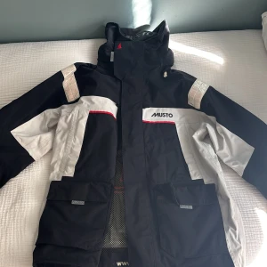 Svart och vit seglarjacka från Musto - Säljer en svart och vit seglarjacka från Musto med huva och reflekterande detaljer på axlarna. Jackan har flera fickor framtill, dragkedja och justerbar huva. Perfekt för blåsiga dagar på sjön eller utomhusaktiviteter.
